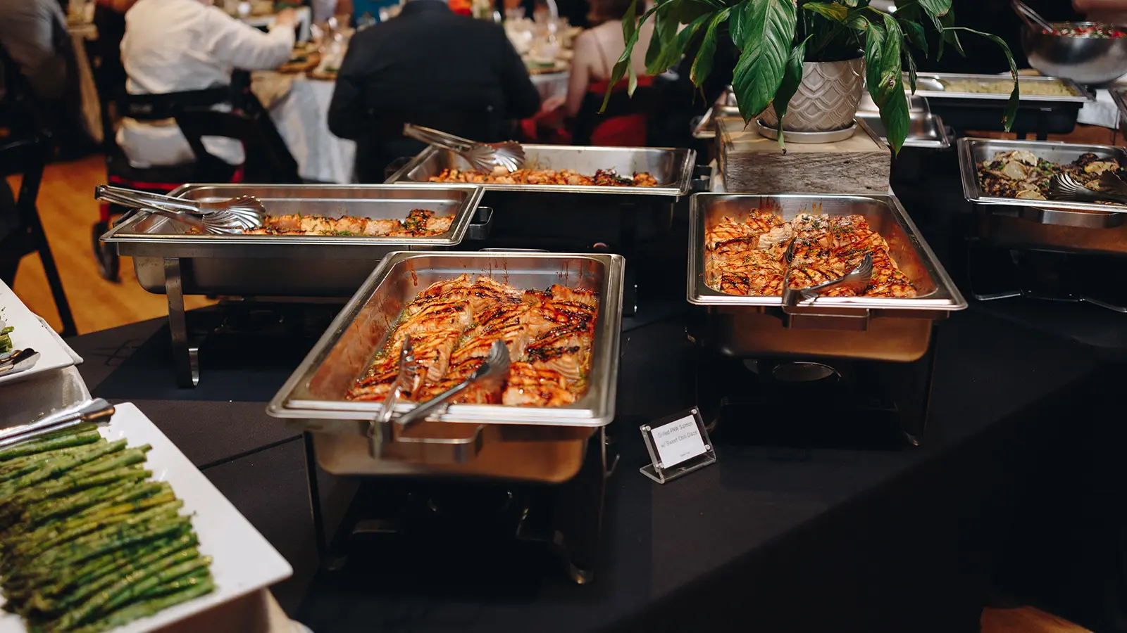 Chafing dishes of grilled salmon in sweet chili glaze and a platter of roasted asparagus on a catering station, with guests seated at tables behind.