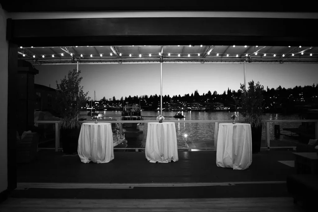 Black-and-white photo of three cocktail tables under string lights on the covered patio at dusk, with the marina and Lake Union behind them.