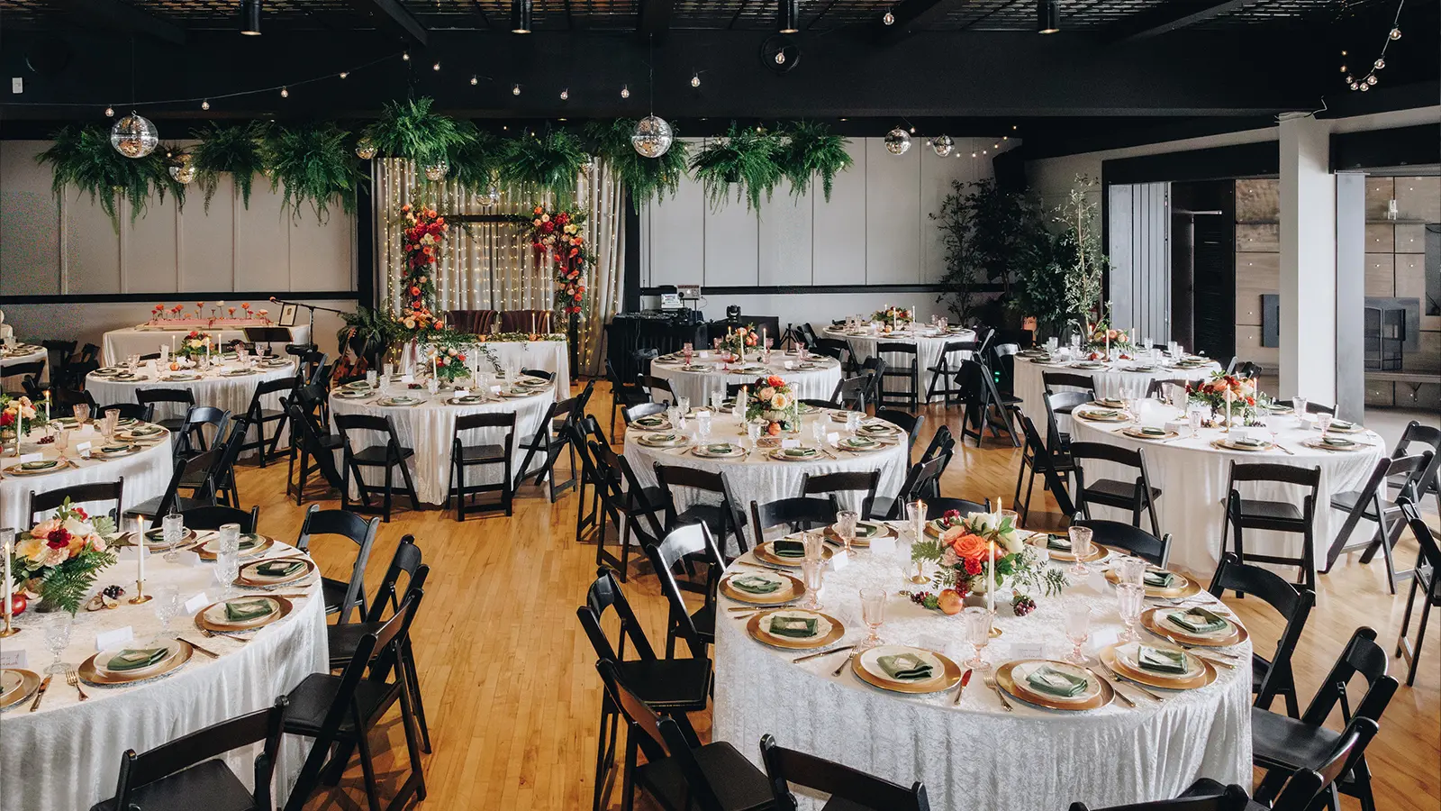 The main room set at rounds for a wedding reception, with a floral backdrop behind the sweetheart table and hanging ferns above the dance floor.